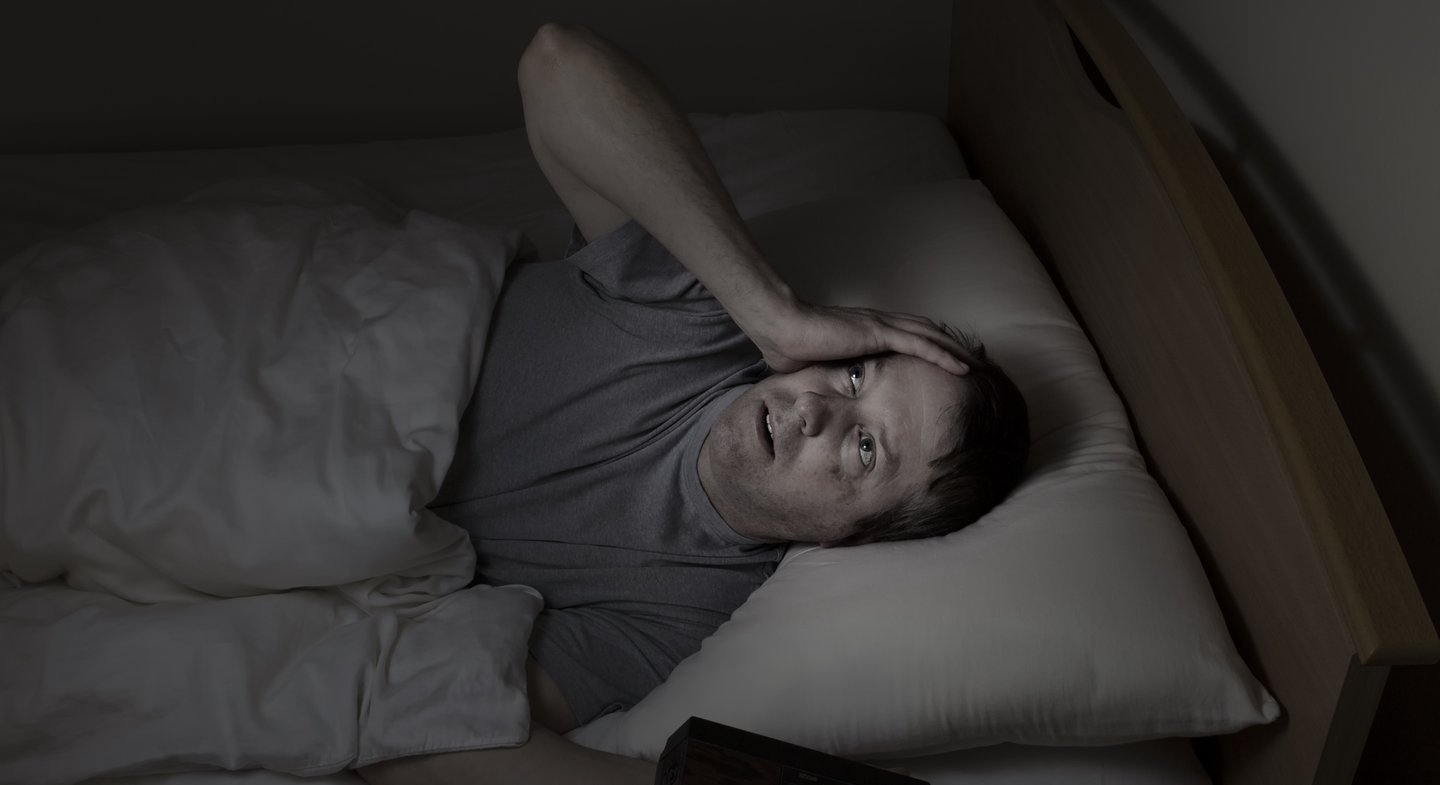 man in darkness staring at ceiling looking troubled, hand on side of face