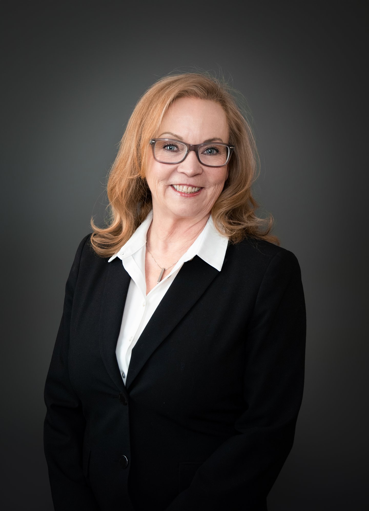 A white woman with red hair and glasses dressed in a black blazer with a while shirt