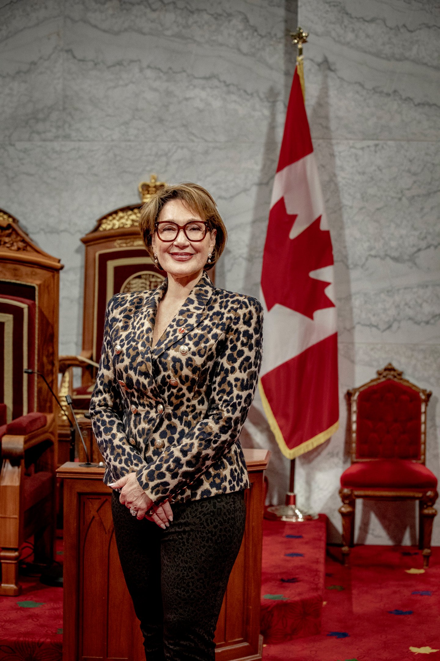Senator Daniele Henkel wearing a leopard print blazer and black plants stands in the senate chamber by a Canadian flag.