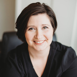 a Caucasian woman with short dark hair smiles at the camera. She is wearing a dark sweater