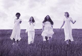 four women of various ages wearing white dresses walk across an open field